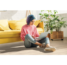 Une femme est assise par terre et utilise un ordinateur portable, avec un canapé et des plantes en arrière-plan.