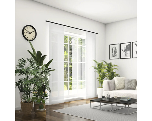 Helles Wohnzimmer mit Sofa, Couchtisch, Zimmerpflanzen, Uhr und Schiebevorhang vor dem Fenster.