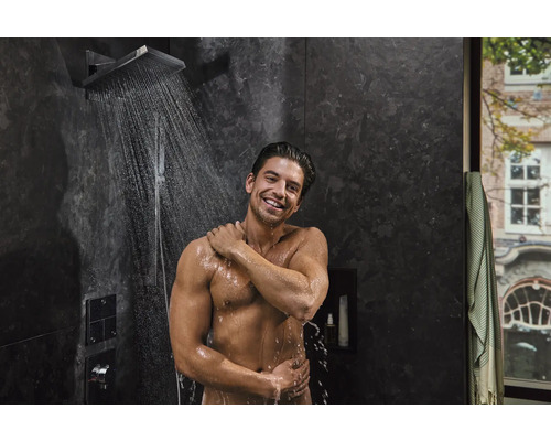 Homme sous un pommeau de douche dans une salle de bains avec des carreaux sombres