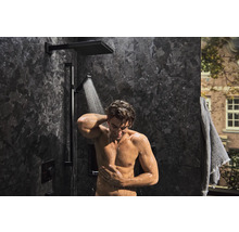 Homme sous une robinetterie de douche moderne avec douche de tête et douchette à main dans une salle de bains avec des carreaux sombres