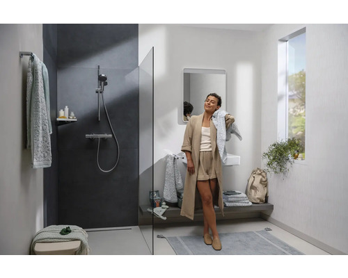 Helles Badezimmer mit Dusche, Waschbecken, Spiegel, Fenster und einer Frau, die sich mit einem Handtuch abtrocknet.