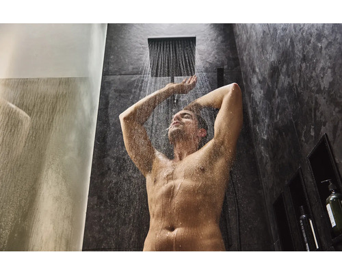 Mann duscht unter einer Regendusche in einem Badezimmer mit dunklen Fliesen.