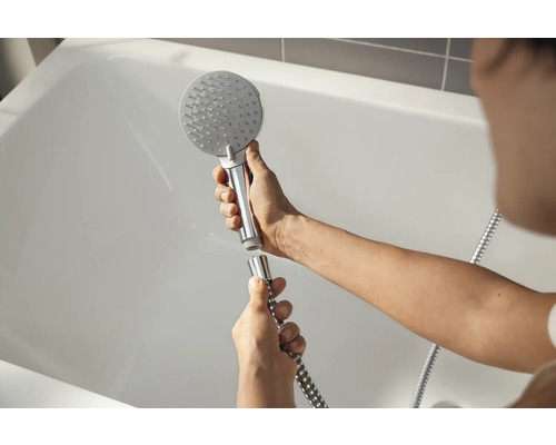 Une personne fixe une pomme de douche sur un flexible de douche devant une baignoire.