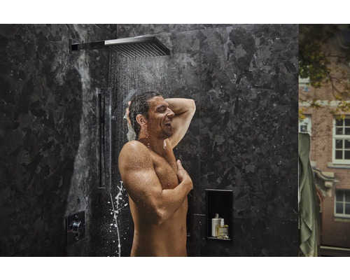 Homme se douche sous une douche de tête dans une salle de bains moderne avec des carreaux foncés.