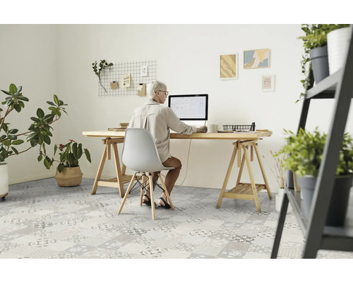 Une femme est assise à un bureau avec un ordinateur dans une pièce lumineuse avec des plantes et un revêtement de sol à motifs.