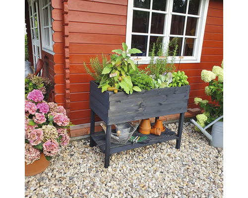 Jardinière en bois avec herbes aromatiques, étagère et outils de jardin devant une maison en bois rouge.