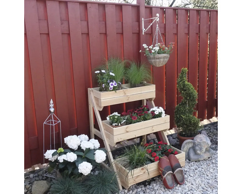 Holzpflanzenständer mit drei Ebenen, bepflanzt mit verschiedenen Blumen und Pflanzen vor einem roten Gartenzaun.