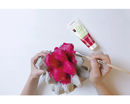 Une personne peint un emballage à œufs avec un pinceau et de la peinture rouge, avec un tube de peinture en arrière-plan.