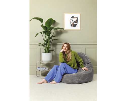 Frau sitzt in einem Zimmer auf einem grauen Sitzsack neben einer Zimmerpflanze und einem Beistelltisch