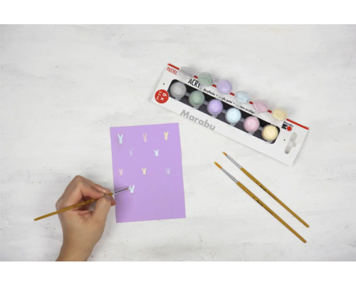 Szene mit einer Person, die mit einem Pinsel und einem Farbkasten auf lila Papier Hasen malt.