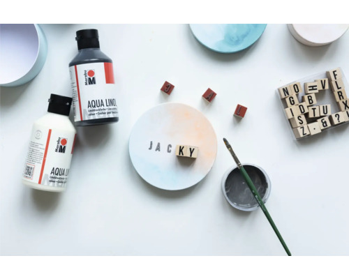 Szene mit Aqua Linoldruckfarbe von Marabu, Pinsel, Holzstempeln und einem beschrifteten Untersetzer mit dem Namen Jacky