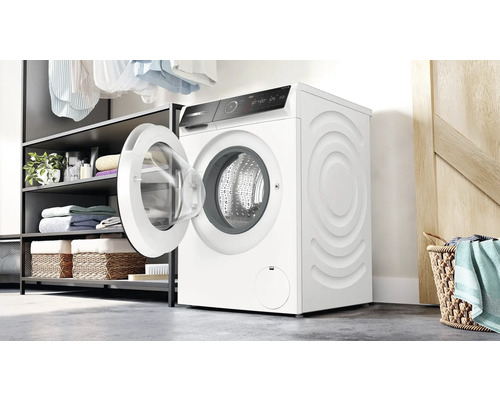 Machine à laver avec porte ouverte dans une buanderie lumineuse, à côté d''une étagère avec du linge et des paniers