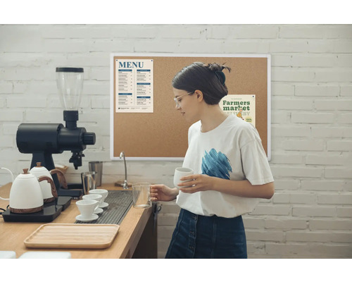 Dans un café lumineux, une femme portant des lunettes se tient derrière le comptoir, tenant une tasse et un verre doseur, entourée d''une machine à café, de bouilloires et d''un tableau de menu sur un mur de briques.