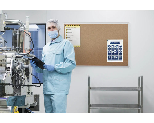 Homme en vêtements de protection avec tablette dans un hall de production, panneaux d''avertissement au mur