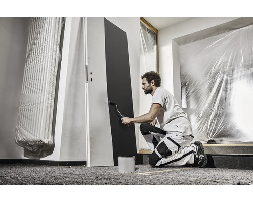 Ein Mann streicht eine Tür mit einer Farbrolle in einem Raum, der mit Schutzfolie abgedeckt ist. Malerarbeiten