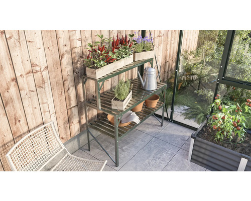 Scène de jardin avec une table de plantation en métal, des caisses en bois plantées, un arrosoir et des outils de jardin dans une serre