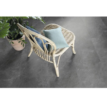 Scène avec une chaise en rotin avec des coussins et une plante en pot sur un revêtement de sol gris.