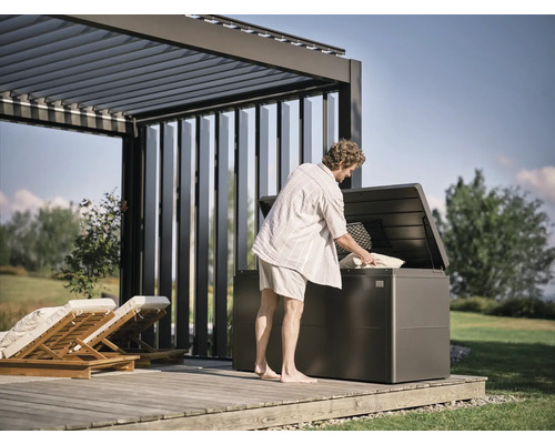 Mann befüllt eine Gartenbox unter einer Pergola auf einer Holzterrasse mit Kissen.