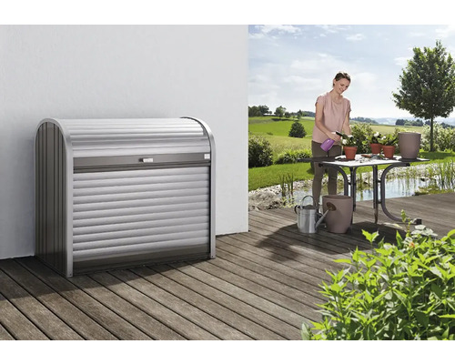Mülltonnenbox Biohort StoreMax 120, 117 x 73 x 109 cm, silber-quarzgrau Szene mit einem Rolladenschrank auf einer Terrasse und einer Frau, die Pflanzen gießt