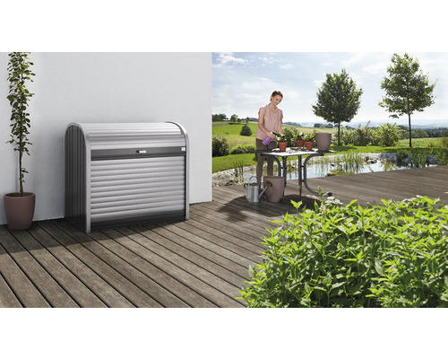 Mülltonnenbox Biohort StoreMax 120, 117x73x109 cm dunkelgrau-metallic Szene mit einem Mülltonnenbox-Design auf einer Holzterrasse und einer Frau, die im Garten Blumen gießt