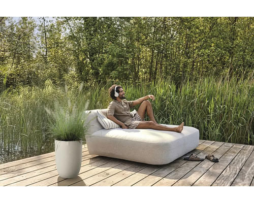 Mann entspannt auf einer Gartenliege mit Kopfhörern, neben ihm ein Pflanzgefäß mit Ziergras auf einer Holzterrasse.