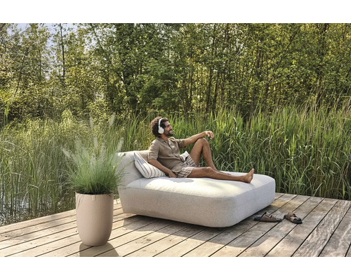 Szene mit Mann auf einer Gartenliege mit Kissen, Topfpflanze und Schuhen auf einer Holzterrasse am See