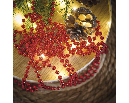 Décoration avec collier de perles rouges, branches de sapin, guirlande lumineuse et pommes de pin sur une assiette