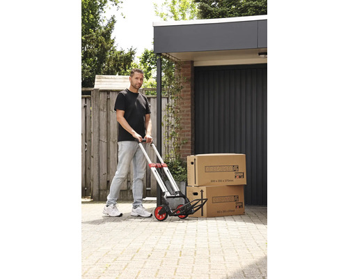 Un homme pousse un diable avec deux cartons de déménagement devant un garage.