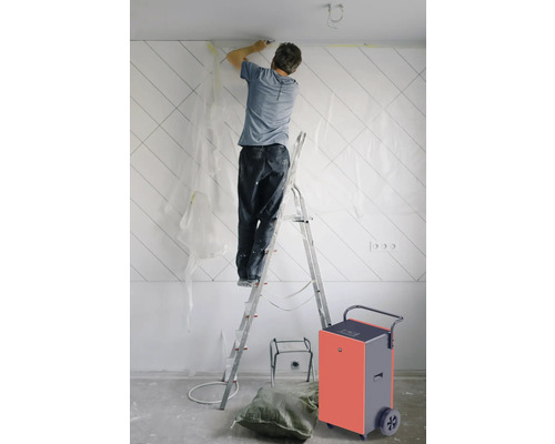 Un homme sur une échelle effectuant des travaux de rénovation avec un déshumidificateur de chantier dans la pièce.