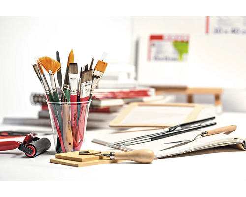 Divers pinceaux dans un gobelet, couteaux à peindre, rouleau à peinture et autres accessoires de peinture sur une table.