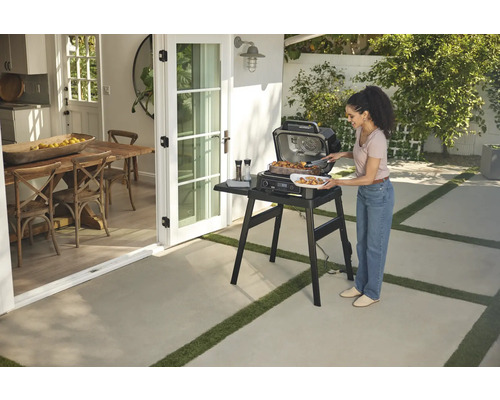 Femme faisant un barbecue avec un gril électrique Ninja à l''extérieur sur une terrasse à côté d''une porte ouverte donnant sur la salle à manger.