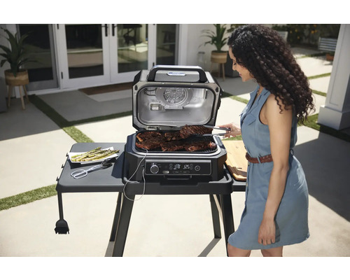 Scène avec une femme faisant griller des steaks sur un gril électrique extérieur, à côté d''une assiette d''asperges.