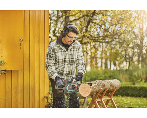 Un homme scie une bûche sur un chevalet à l'aide d'une tronçonneuse à batterie, tout en portant des lunettes de protection et un casque antibruit.