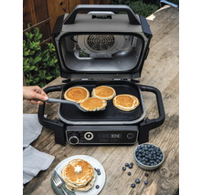Gril Ninja ouvert avec des crêpes sur une plaque de cuisson et une assiette de myrtilles sur une table en bois.