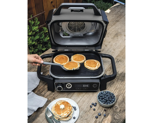 Offener Ninja Grill mit Pfannkuchen auf einer Grillplatte und einem Teller mit Blaubeeren auf einem Holztisch.