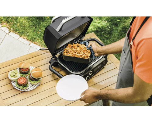 Szene mit einem offenen Elektrogrill, zubereiteten Burgern und Pommes auf einem Gartentisch.