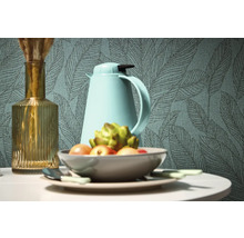 Nature morte décorative avec vase, thermos et coupe de fruits devant un papier peint à motifs de feuilles.