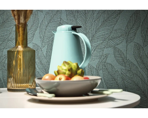 Nature morte décorative avec vase, thermos et coupe de fruits devant un papier peint à motifs de feuilles.