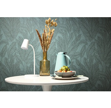 Table à manger décorative avec lampe, thermos, vase avec fleurs séchées et coupe de fruits devant un papier peint à motifs de feuilles