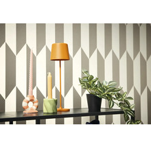 Dekorative Szene mit Lampe, Kerzen und Topfpflanze auf einem Regal vor einer geometrischen Tapete.