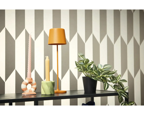 Dekorative Szene mit Lampe, Kerzen und Topfpflanze auf einem Regal vor einer geometrischen Tapete.