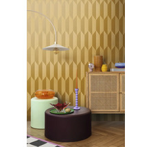 Einrichtungsbeispiel mit einer Lampe, verschiedenen Tischen und einem Schrank vor einer Tapete mit geometrischem Muster.