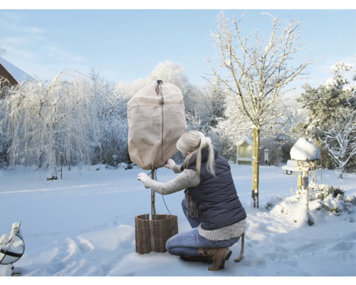 Femme protégeant un arbre avec une housse de protection hivernale dans un jardin enneigé.