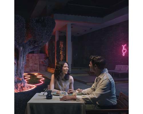 Un couple est assis à une table dans le jardin la nuit, éclairé par des rubans lumineux flexibles et une décoration en forme de cœur au néon.