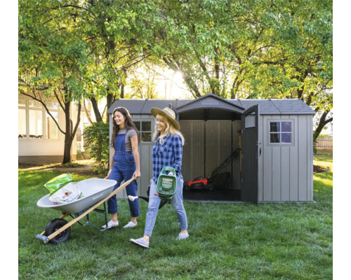 Gartenszene mit zwei Frauen, die eine Schubkarre und einen Rasensprenger tragen, im Hintergrund ein Gartenschuppen