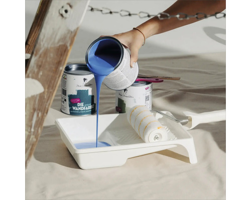 Blaue Wandfarbe wird in eine Farbwanne gegossen, daneben stehen Farbdosen und eine Farbwalze.