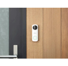 Sonnette vidéo Imou blanche avec caméra et bouton d'appel sur un mur en bois clair à côté d'une porte sombre moderne avec poignée en métal.