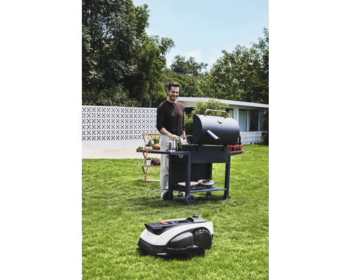 Mann grillt im Garten mit einem Grillwagen und einem Rasenmähroboter.