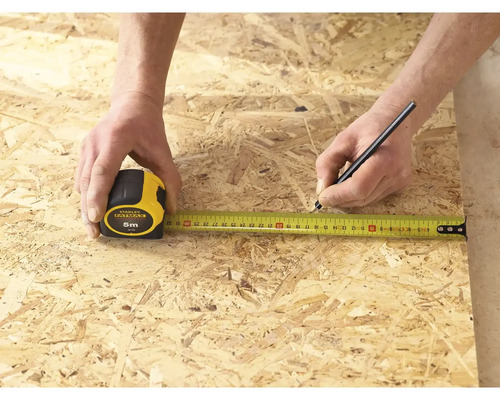 Une personne mesure un morceau de bois avec un mètre ruban Stanley Fatmax de 5 mètres et le marque avec un crayon.