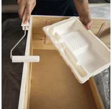 Eine Person streicht ein Holzmöbelstück mit einem Farbroller und einer Farbwanne.